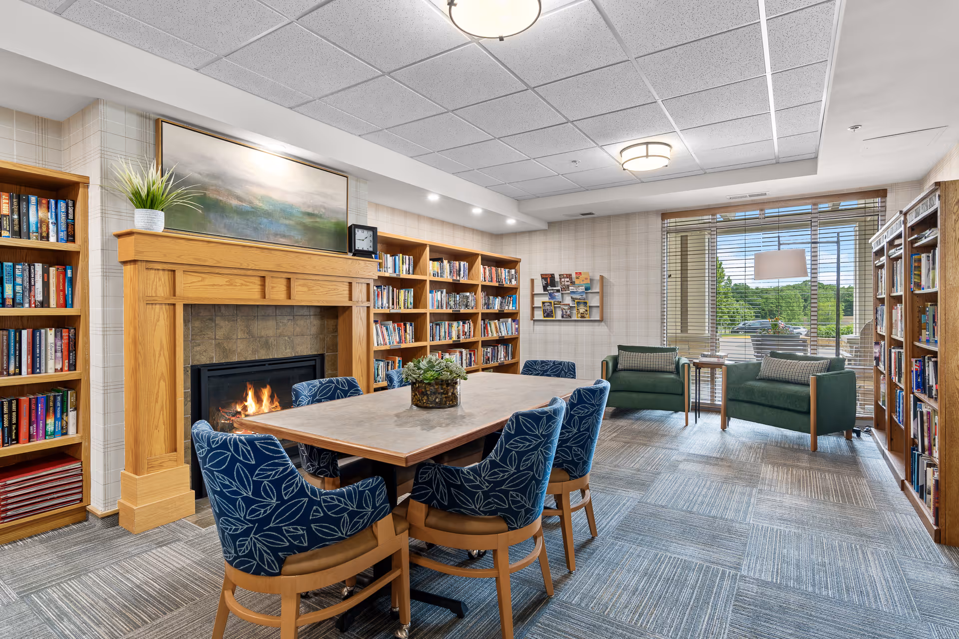 A cozy library room with a wooden fireplace, a table surrounded by six blue patterned chairs, bookshelves filled with books, two green armchairs near a large window with blinds, and a small side table between the armchairs. The room has a carpeted floor and ceiling lights.