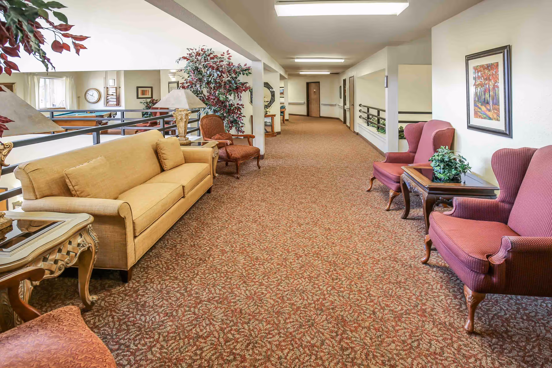 A spacious hallway in a senior living facility with patterned carpet, a beige sofa, red upholstered armchairs, side tables with lamps and plants, framed artwork on the walls, and a railing overlooking a lower level with a pool table and clock on the wall.