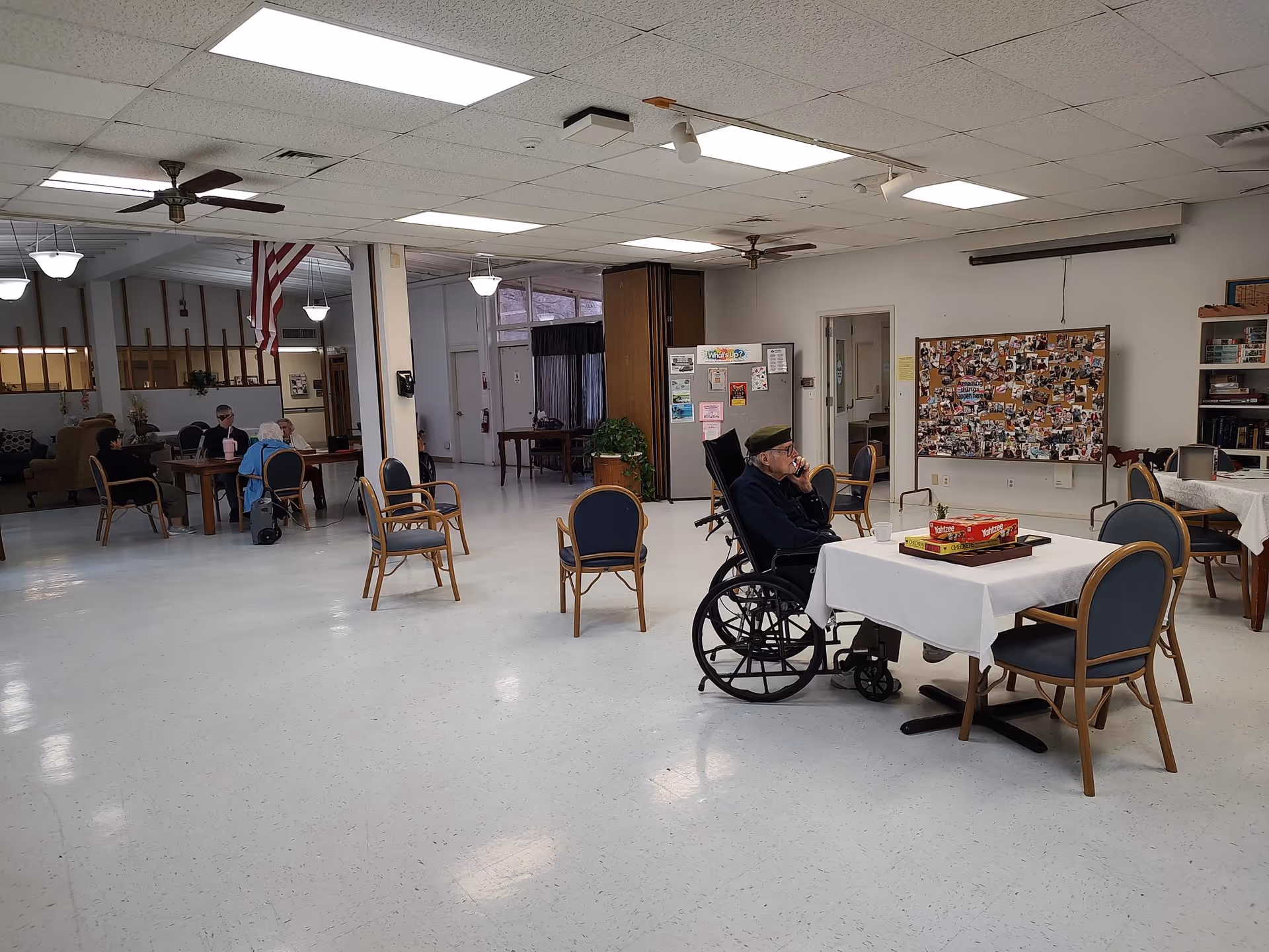 A spacious common area in an assisted living facility with several tables and chairs. An elderly man in a wheelchair sits at a table with a board game on it. In the background, a few other elderly individuals are seated at another table. The room has white walls, a bulletin board filled with photos and notes, ceiling fans, and fluorescent lighting.