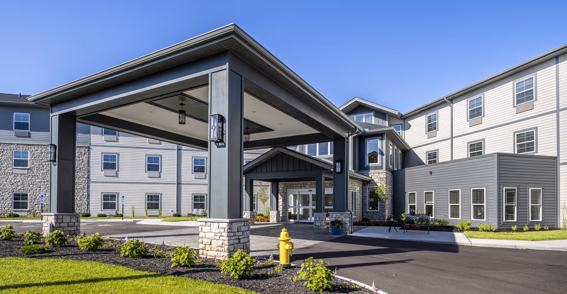 Entrance canopy and porte-cochère of a modern multi-story senior living building with landscaped grounds under a clear blue sky.