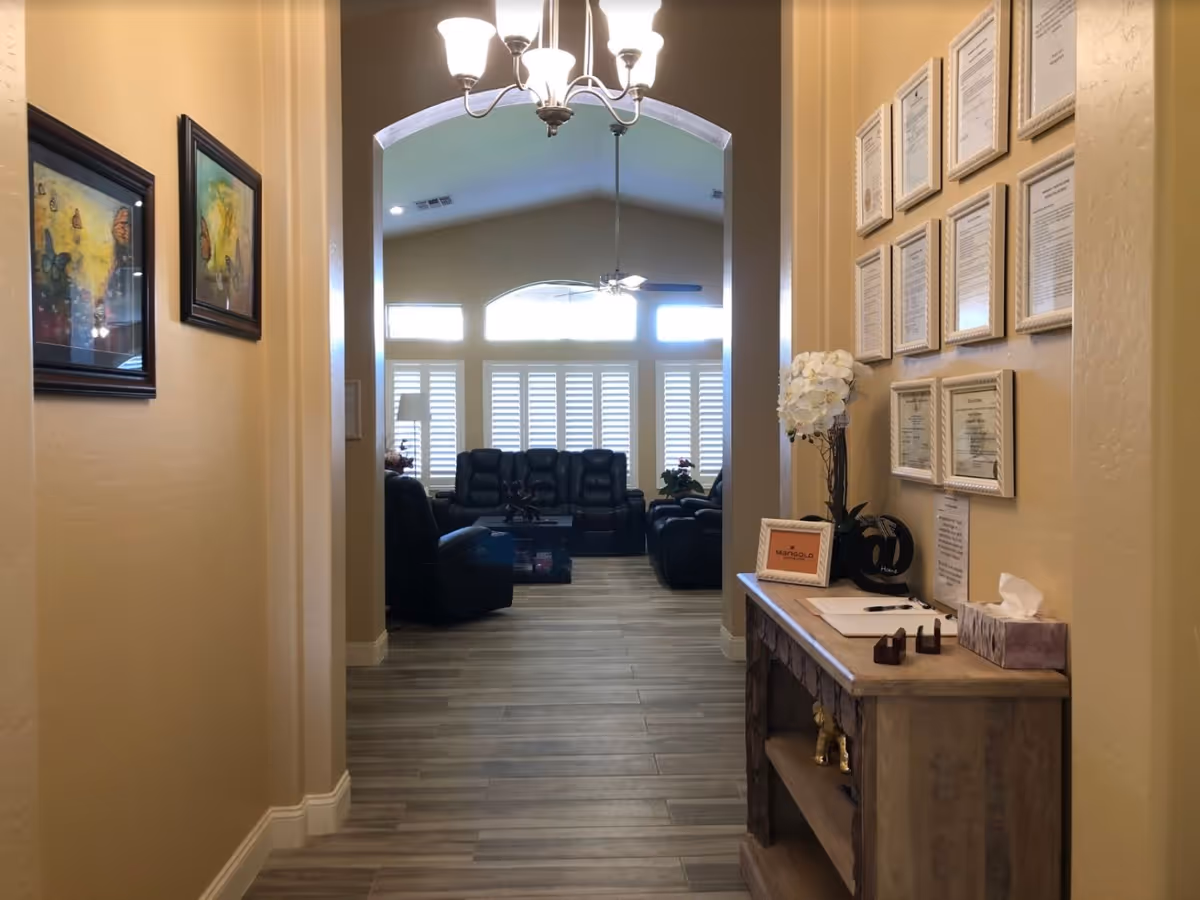 View down a hallway with beige walls and wood-look tile flooring leading to a living room area with black leather recliners and large windows with white shutters. On the right side of the hallway is a wooden console table with framed documents, a flower vase, and a tissue box. Two framed butterfly pictures hang on the left wall. A chandelier light fixture hangs from the ceiling.