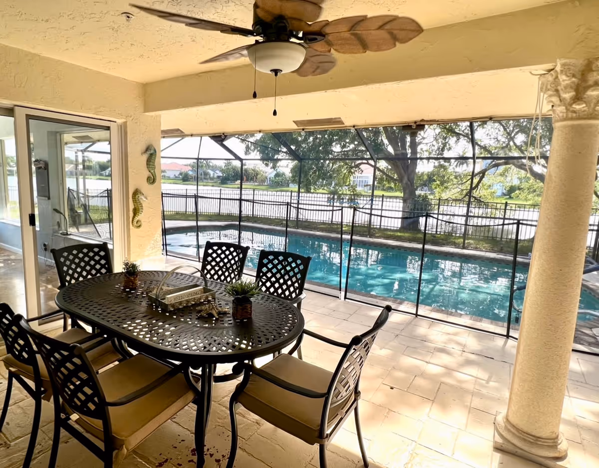 Covered outdoor patio area with a ceiling fan, a black metal table with six matching chairs, and decorative items on the table. Beyond the patio is a fenced swimming pool with a view of a lake and trees in the background.