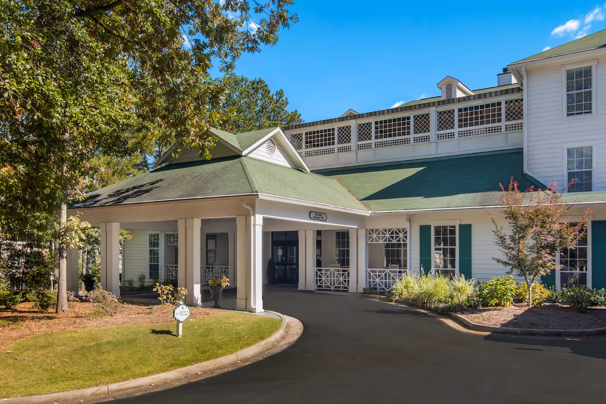 Exterior view of Arbor Terrace Decatur senior living facility entrance with a covered driveway, white building with green roof, surrounded by trees and landscaping under a clear blue sky.