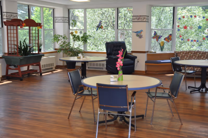 A bright and spacious common area with round tables and chairs arranged on a wooden floor. Large windows let in natural light and show green trees outside. The room features a black recliner chair, potted plants, and colorful wall decorations including birds and butterflies. There is also a cushioned bench along the window.