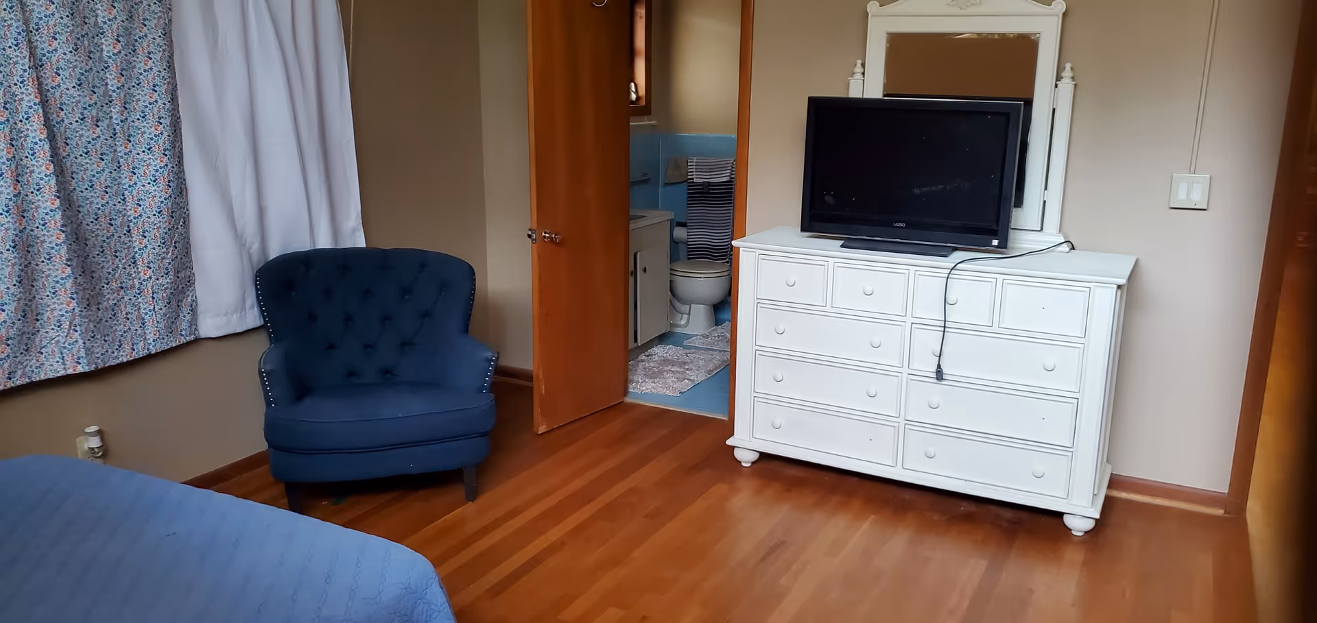 A bedroom with wooden flooring, a blue upholstered armchair, a bed with a blue bedspread, and a white dresser with a TV on top. The door to a small bathroom is open, showing a toilet, sink, and blue tiled walls. There are floral and white curtains on the window.