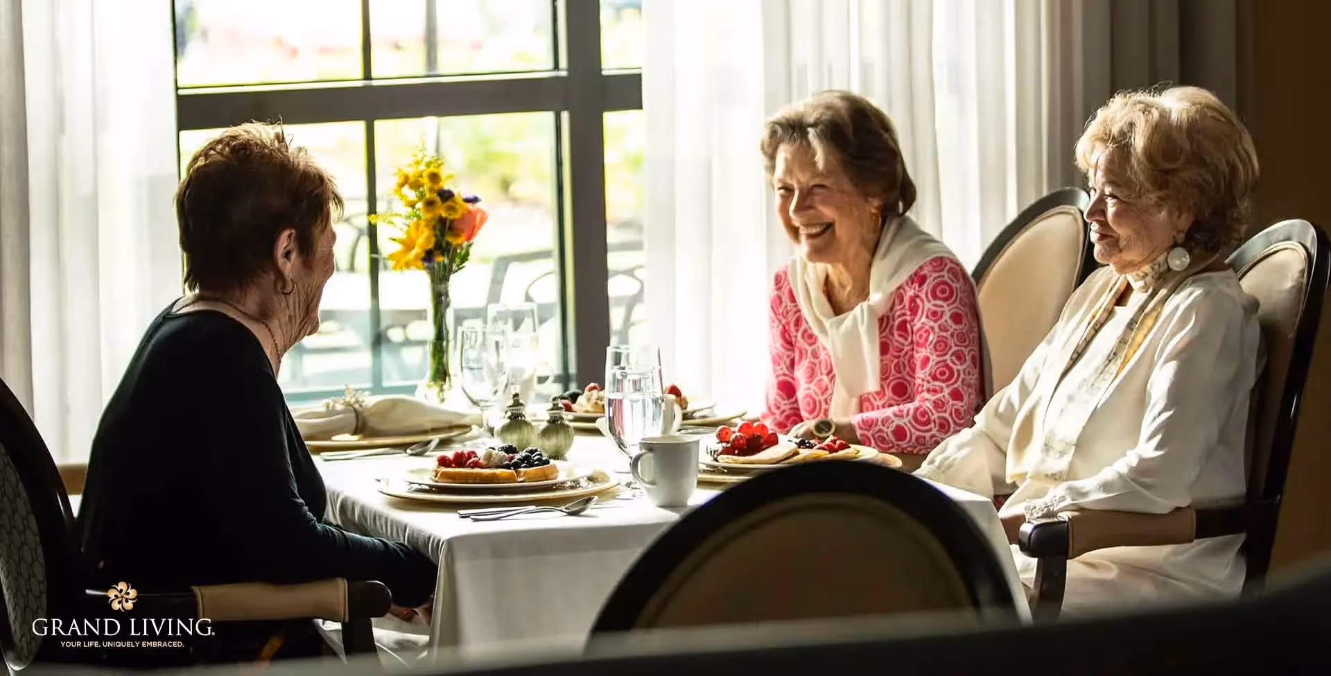 Three elderly women sitting around a dining table with plates of food and glasses of water, engaged in conversation and smiling, with a window and sheer curtains in the background.