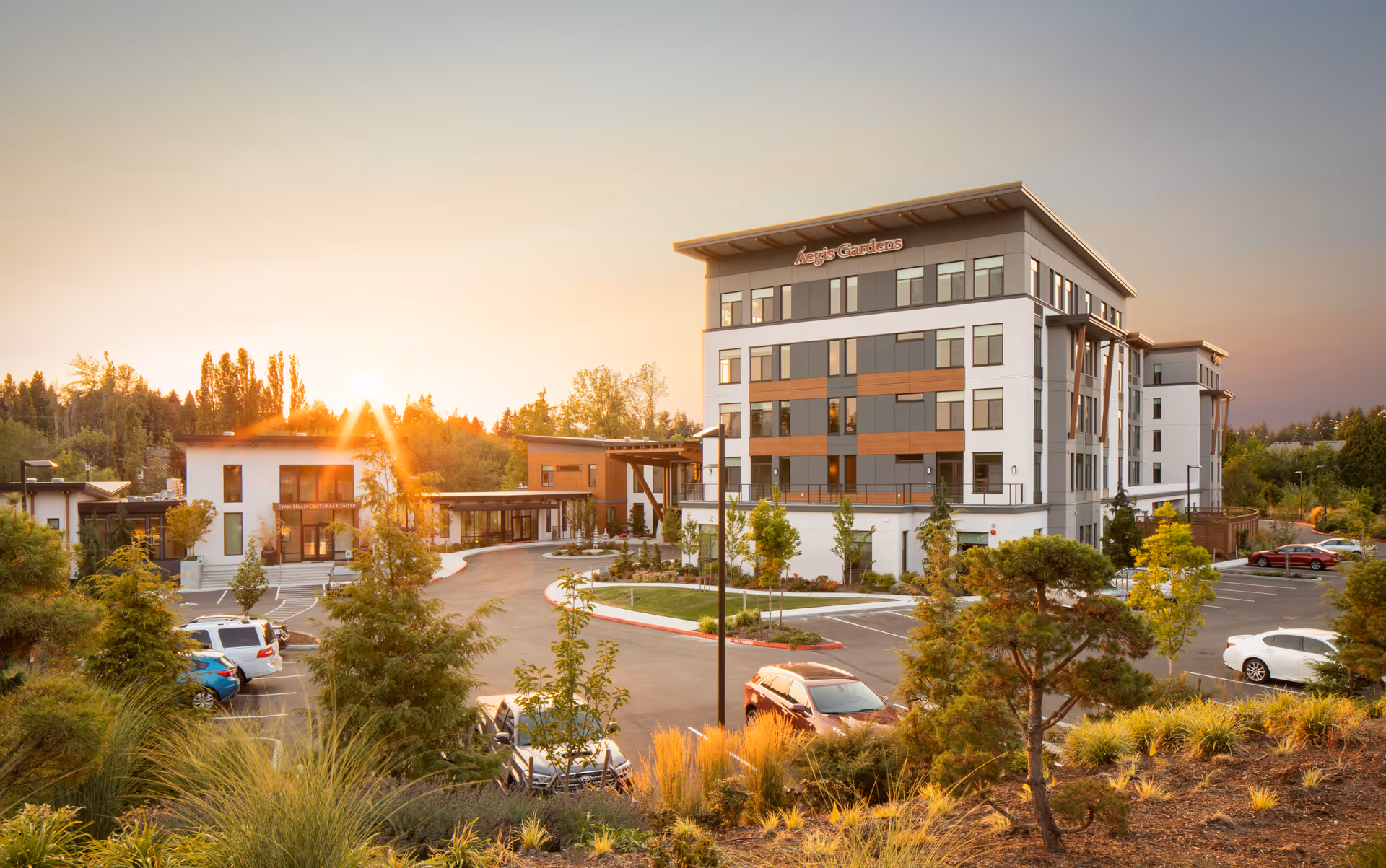 Exterior view of the Aegis Gardens Newcastle senior living building, landscaped grounds and parking lot at sunset.