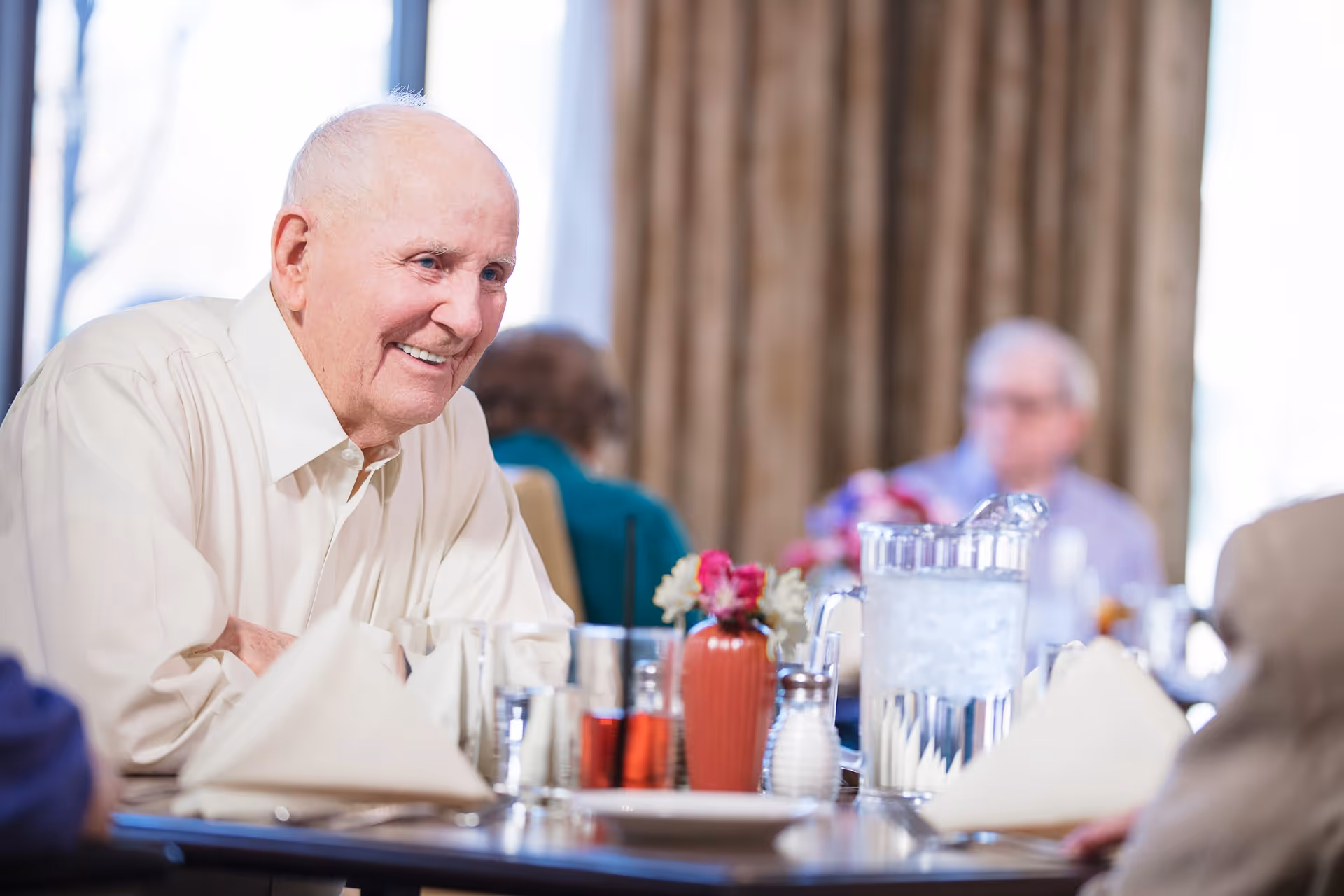 An elderly man smiling and sitting at a dining table set with napkins, glasses, a pitcher of water, and a small vase with flowers in a senior living facility dining room.