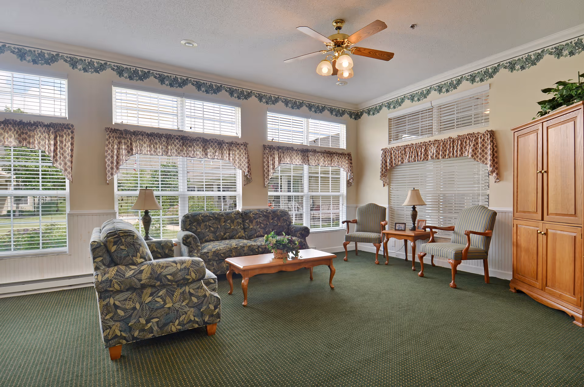 Sunlit common living room with patterned sofa, armchairs, a wooden coffee table and large windows.