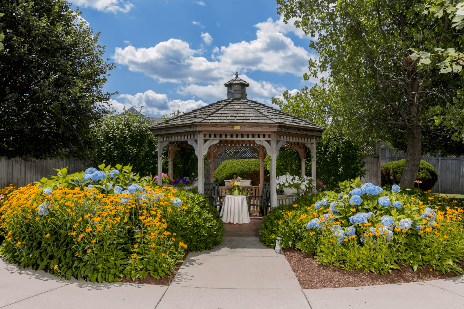 A wooden gazebo surrounded by vibrant flower beds with yellow and blue flowers, set in a garden area with trees and a partly cloudy sky overhead. Inside the gazebo, there is a small round table covered with a white tablecloth and chairs around it.
