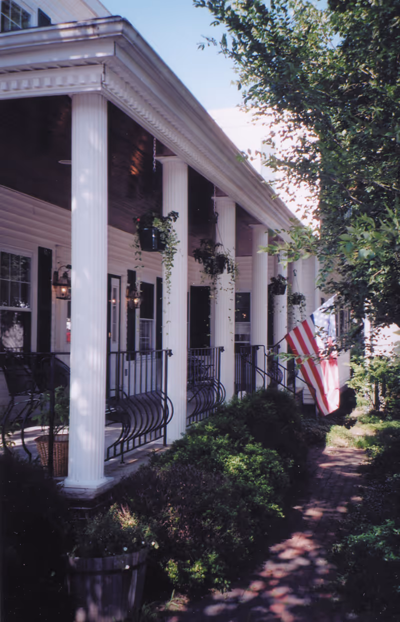 Front porch of a white columned building with hanging plants, railings, and an American flag alongside a brick walkway.