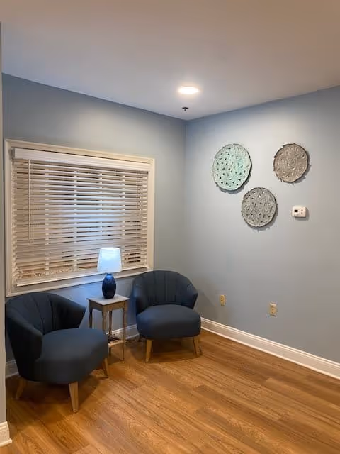 A small seating area with two dark blue upholstered chairs and a wooden side table with a blue lamp on it. The walls are painted light blue, and there are three decorative round wall hangings above a thermostat. The floor is wooden, and there is a window with white blinds.