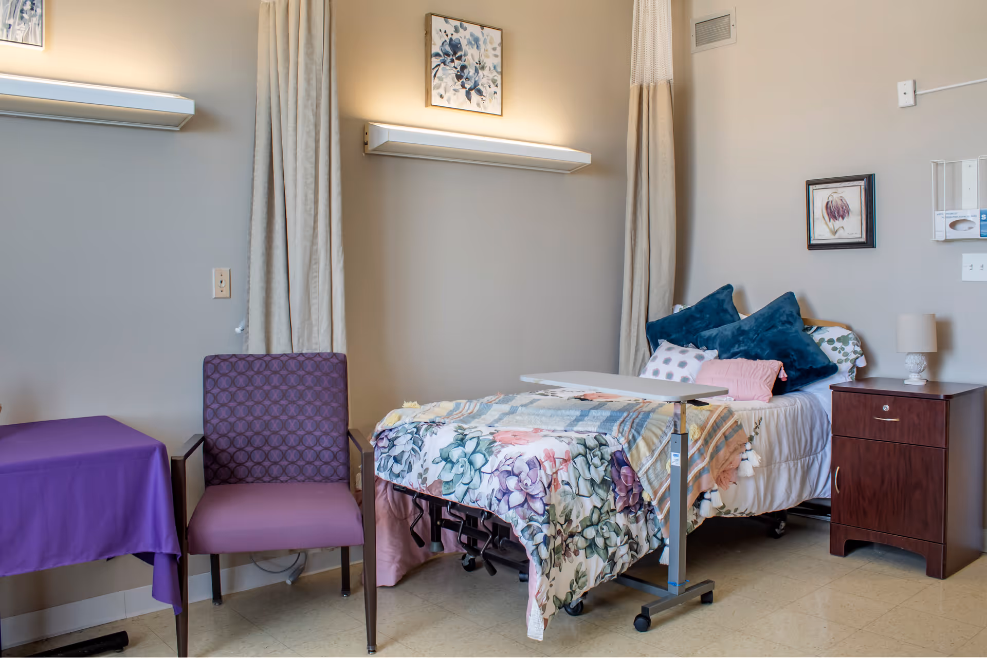 A cozy room in a senior living facility featuring a hospital-style bed with floral bedding and multiple pillows, a wooden nightstand with a lamp, a purple patterned chair, and a small table covered with a purple cloth. The walls are beige with two framed artworks and two wall-mounted lights above the bed area.