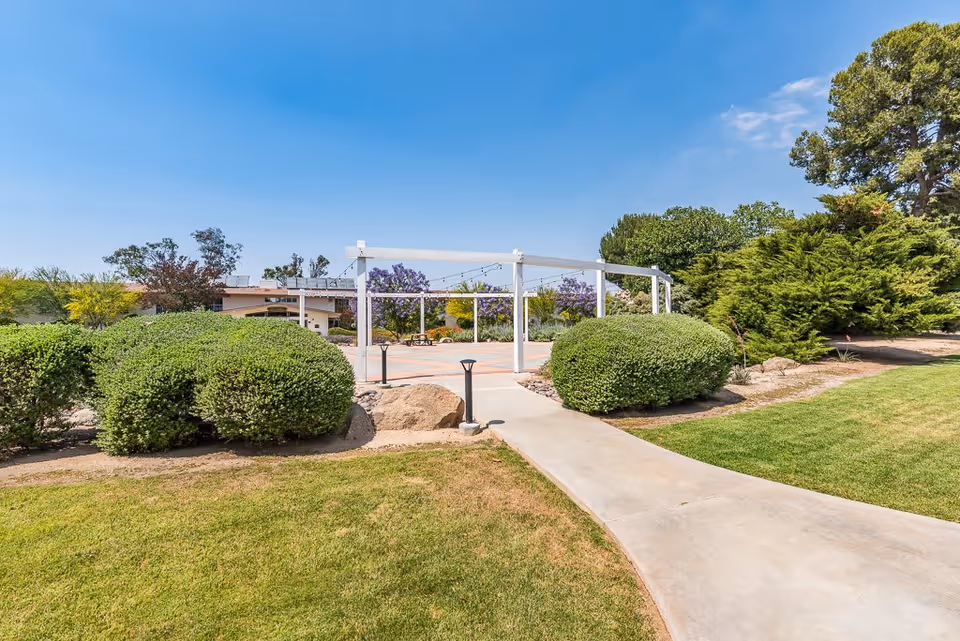 Outdoor garden area with a concrete pathway leading to a white pergola structure surrounded by green bushes, trees, and a well-maintained lawn under a clear blue sky.