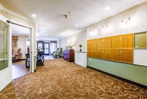Interior hallway of a senior living facility with patterned carpet, a row of yellow mailboxes on the right wall, and a sitting area with chairs and a table visible in the background. There is a doorway on the left leading to a room with armchairs and a fireplace.