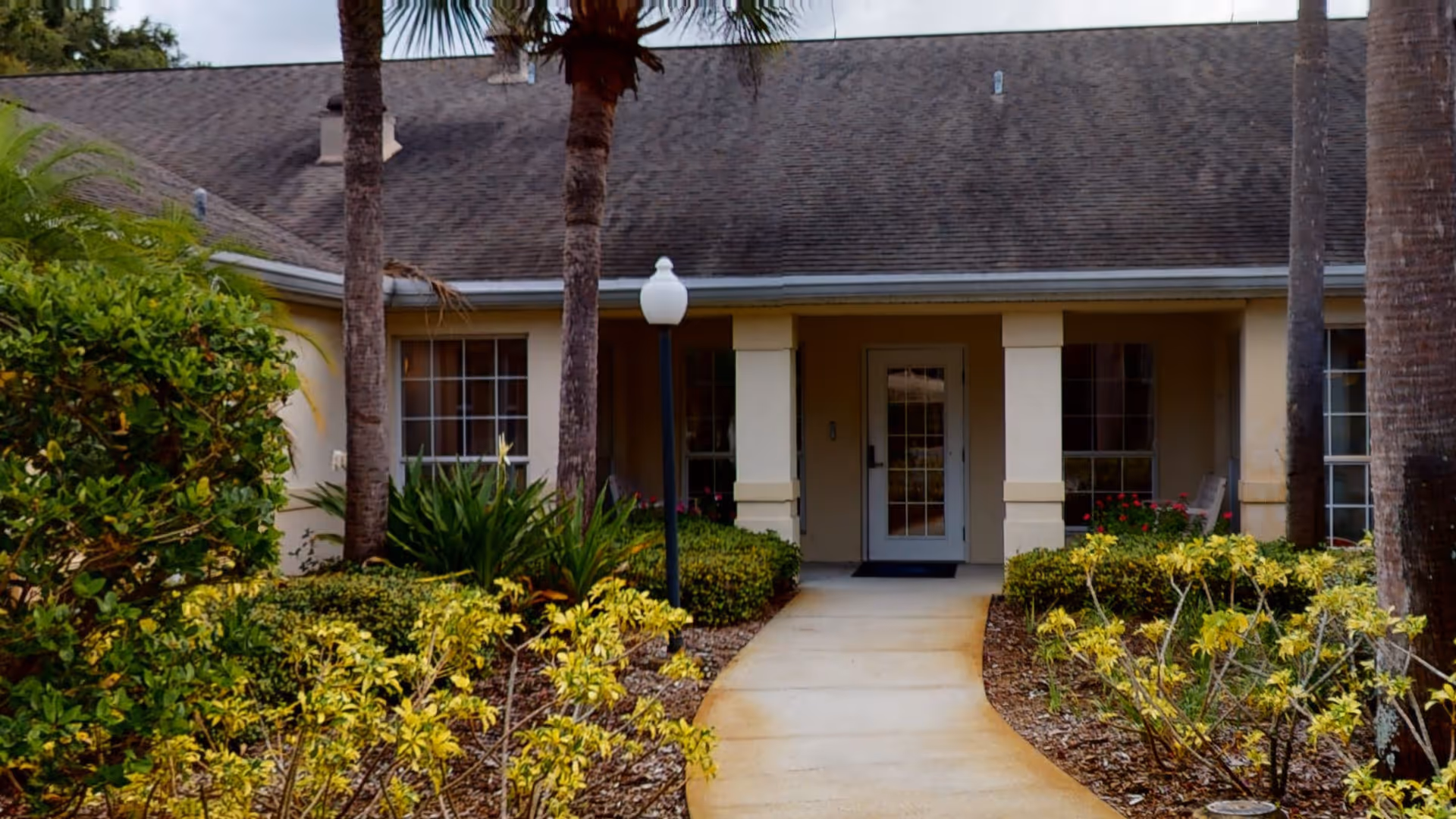 Exterior view of a single-story building with a walkway leading to a glass door entrance. The building is surrounded by landscaped bushes, plants, and palm trees, with a lamp post near the walkway.