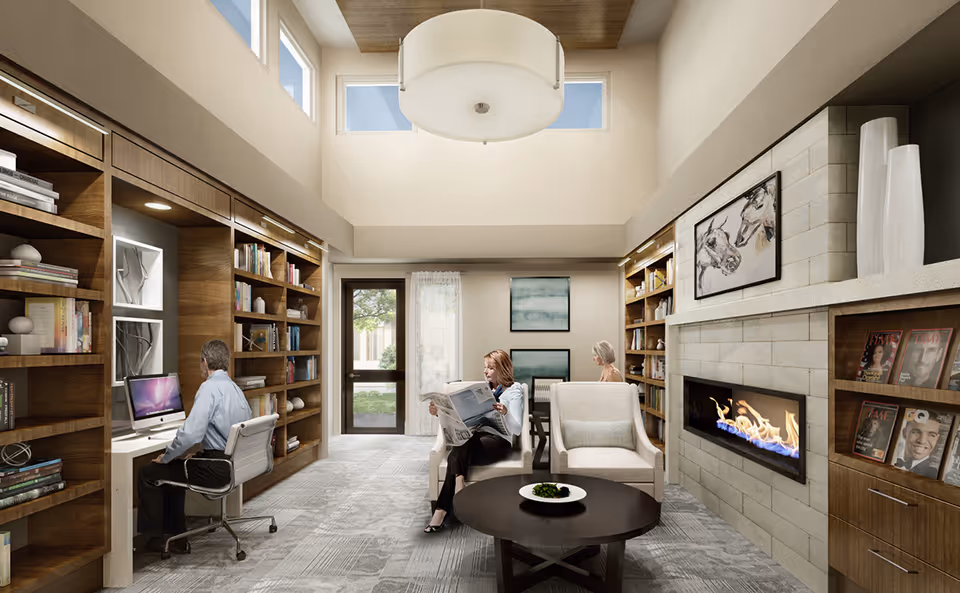 A cozy senior living common area with high ceilings and large windows. Two women are seated on armchairs near a modern fireplace, one reading a newspaper and the other sitting at a table. A man is seated at a built-in desk with a computer on the left side. The room features wooden bookshelves filled with books and decorative items, a round coffee table with a small plant, and framed artwork on the walls.