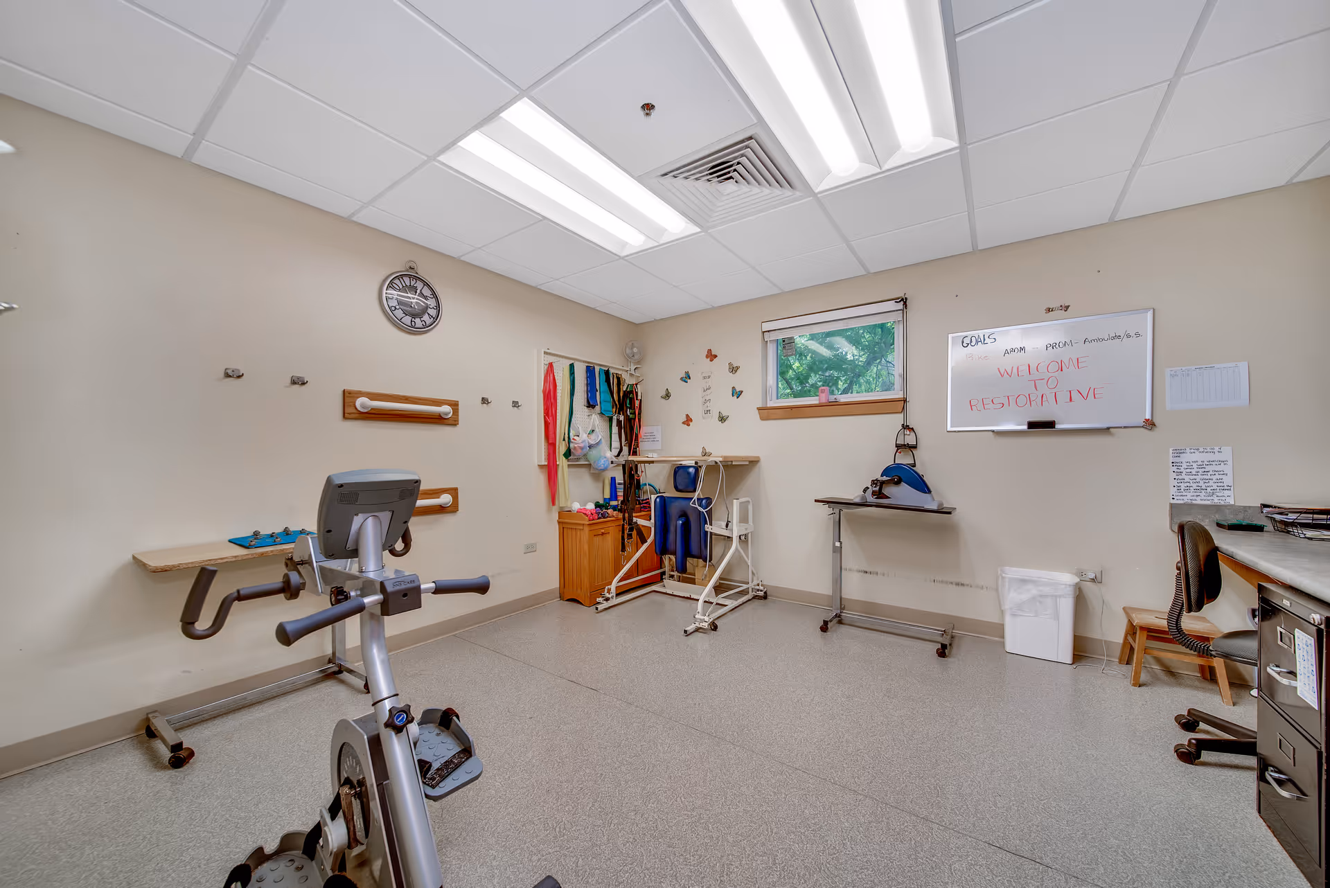 A therapy or exercise room with an exercise bike, various therapy equipment, a desk with a chair, a whiteboard with the message 'WELCOME TO RESTORATIVE', a clock on the wall, and a window showing greenery outside.