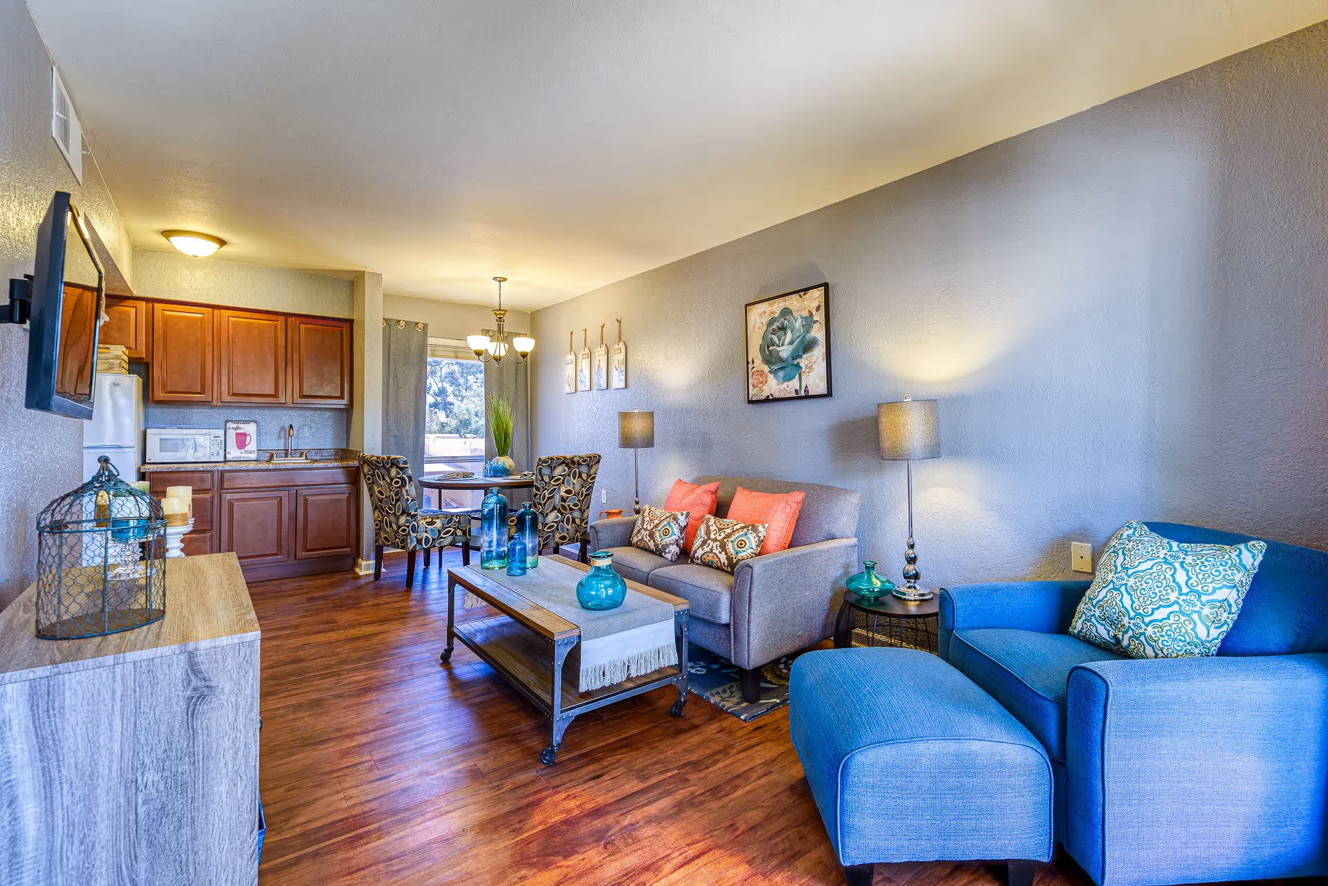 A cozy living room and dining area in a senior living facility with wooden flooring, a gray sofa with colorful cushions, a blue armchair with a matching ottoman, a wooden coffee table with decorative blue glass bottles, a small round side table with a lamp, and a dining table with four patterned chairs near a window. The kitchen area has wooden cabinets, a microwave, and a refrigerator.
