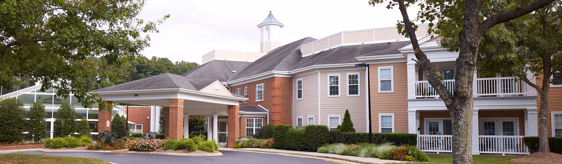 Front entrance of a multi-story senior living facility with a covered porte-cochere, balconies, and landscaped grounds.