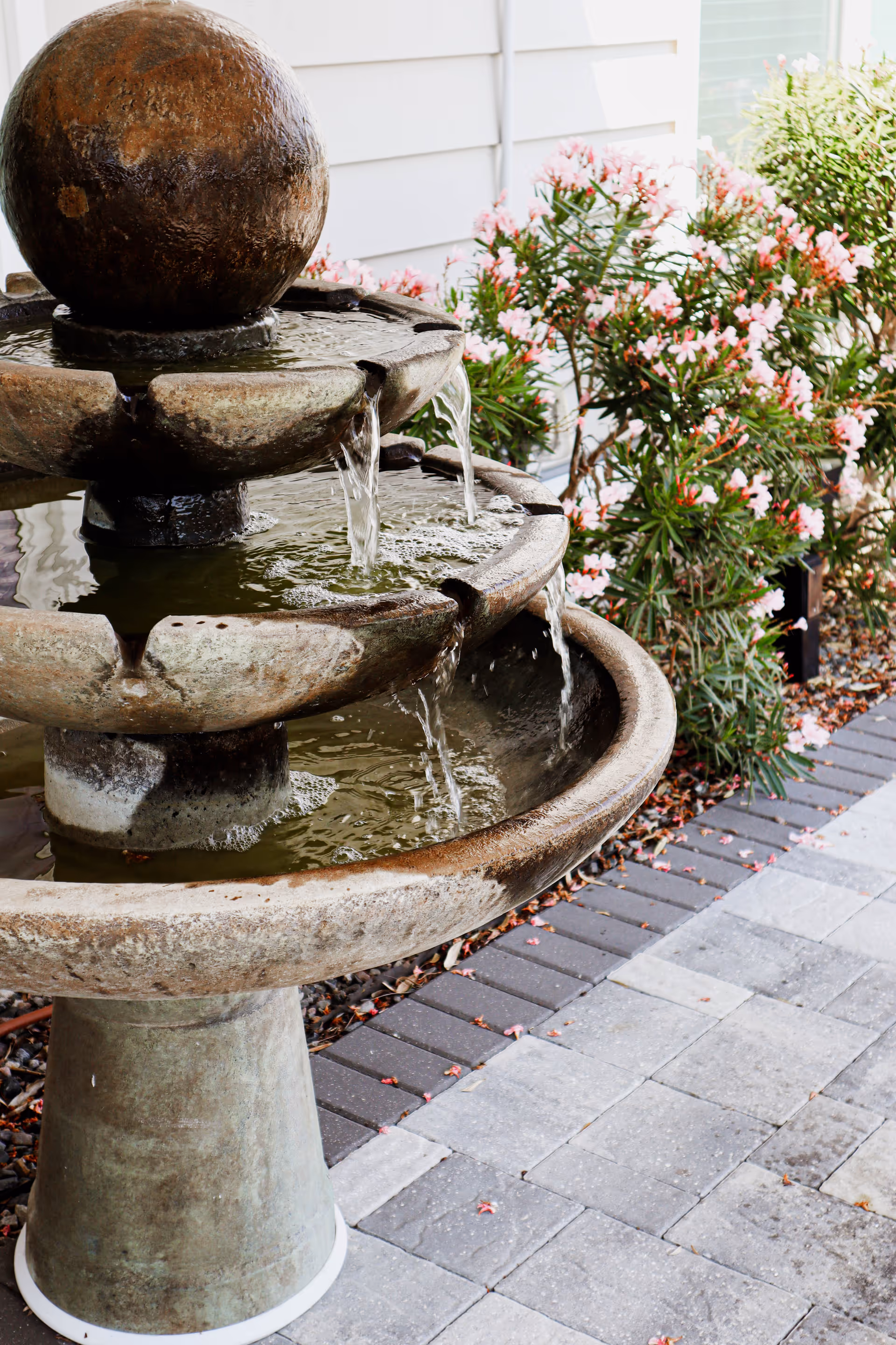 A three-tiered stone water fountain with water flowing from the top spherical element down to the lower basins, situated on a paved area next to a garden with pink flowering bushes and a white building wall in the background.
