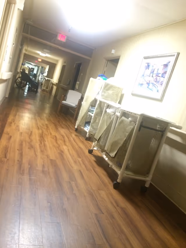 A long hallway in Woodland Care Center with wooden flooring and beige walls. On the right side, there are laundry carts with cloth bags and a framed picture hanging on the wall. A chair is placed against the wall further down the hallway. At the far end, a person in a wheelchair is visible near an exit sign.