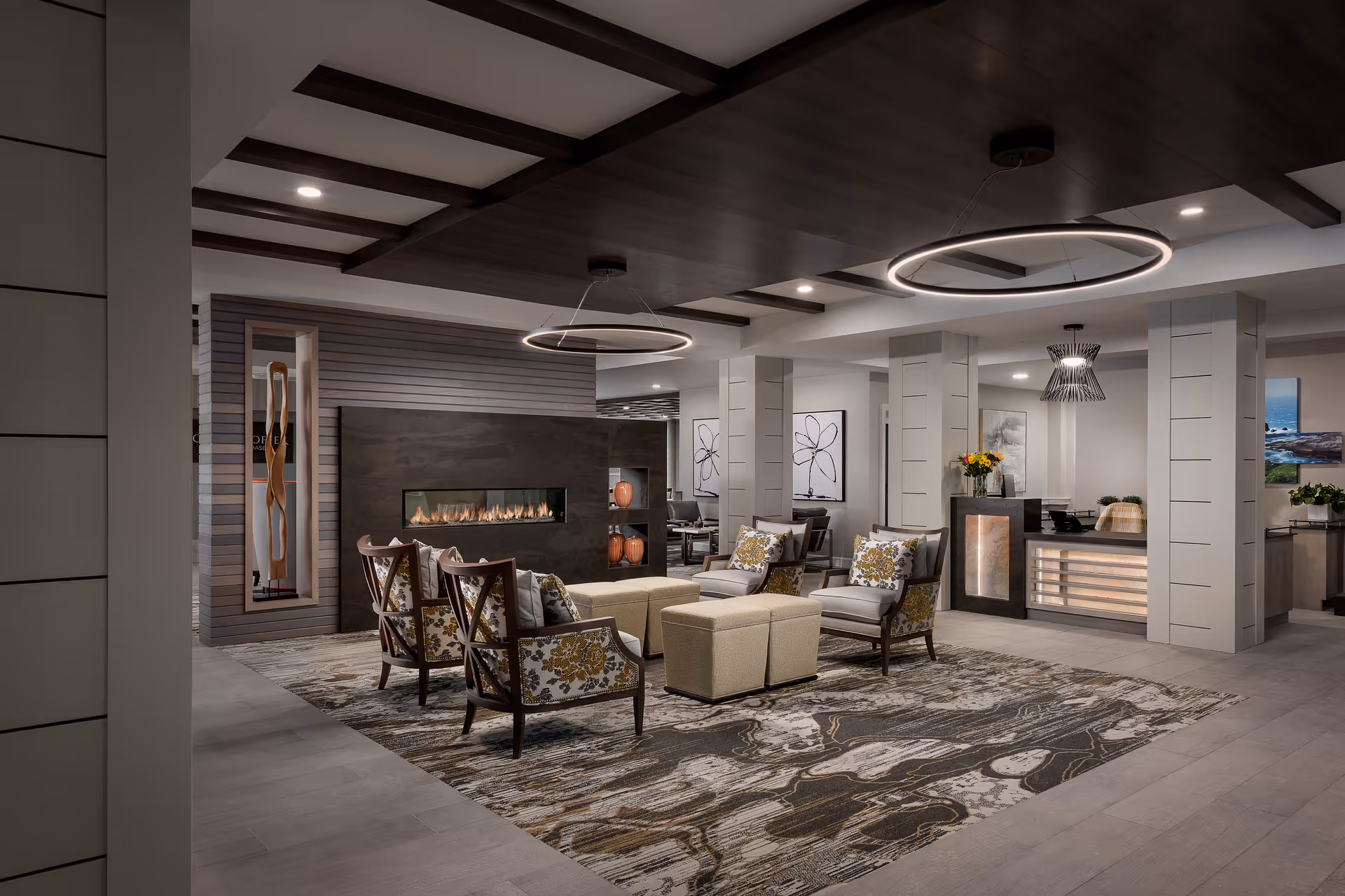 A modern and elegant senior living facility common area with a seating arrangement consisting of four floral-patterned armchairs and two beige ottomans around a rectangular rug. The room features a contemporary linear fireplace set into a dark accent wall with decorative vases, circular pendant lights hanging from a dark wood ceiling panel, and a reception desk in the background with flowers and artwork on the walls.