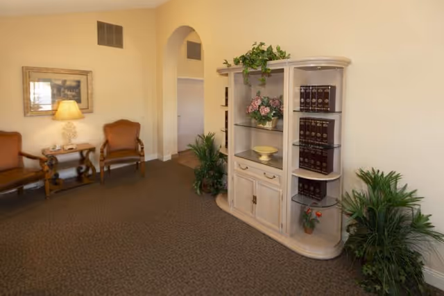 A cozy interior room with two wooden armchairs with brown cushions, a small wooden side table with a lamp and a framed picture on the wall. There is a light-colored wooden cabinet with glass shelves displaying books and decorative plants. The room has beige walls, carpeted floor, and several green potted plants near the cabinet and archway.