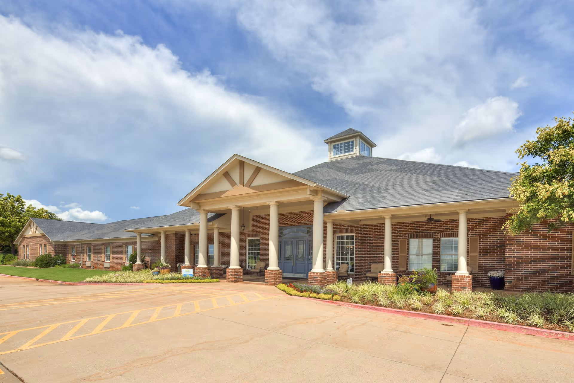 Exterior view of a single-story brick building with a covered entrance supported by white columns, surrounded by landscaping and a clear blue sky with some clouds.