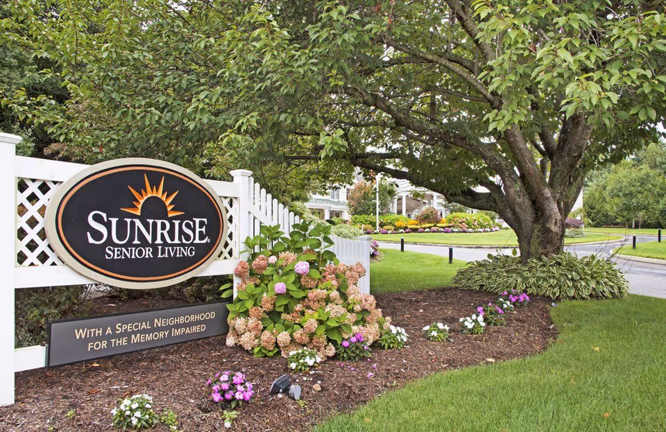 Outdoor view of the entrance sign for Sunrise Senior Living, surrounded by a white lattice fence, flowering plants, and a large tree with green leaves. The sign includes the text 'Sunrise Senior Living' and 'With a Special Neighborhood for the Memory Impaired'. A paved road and landscaped grounds are visible in the background.