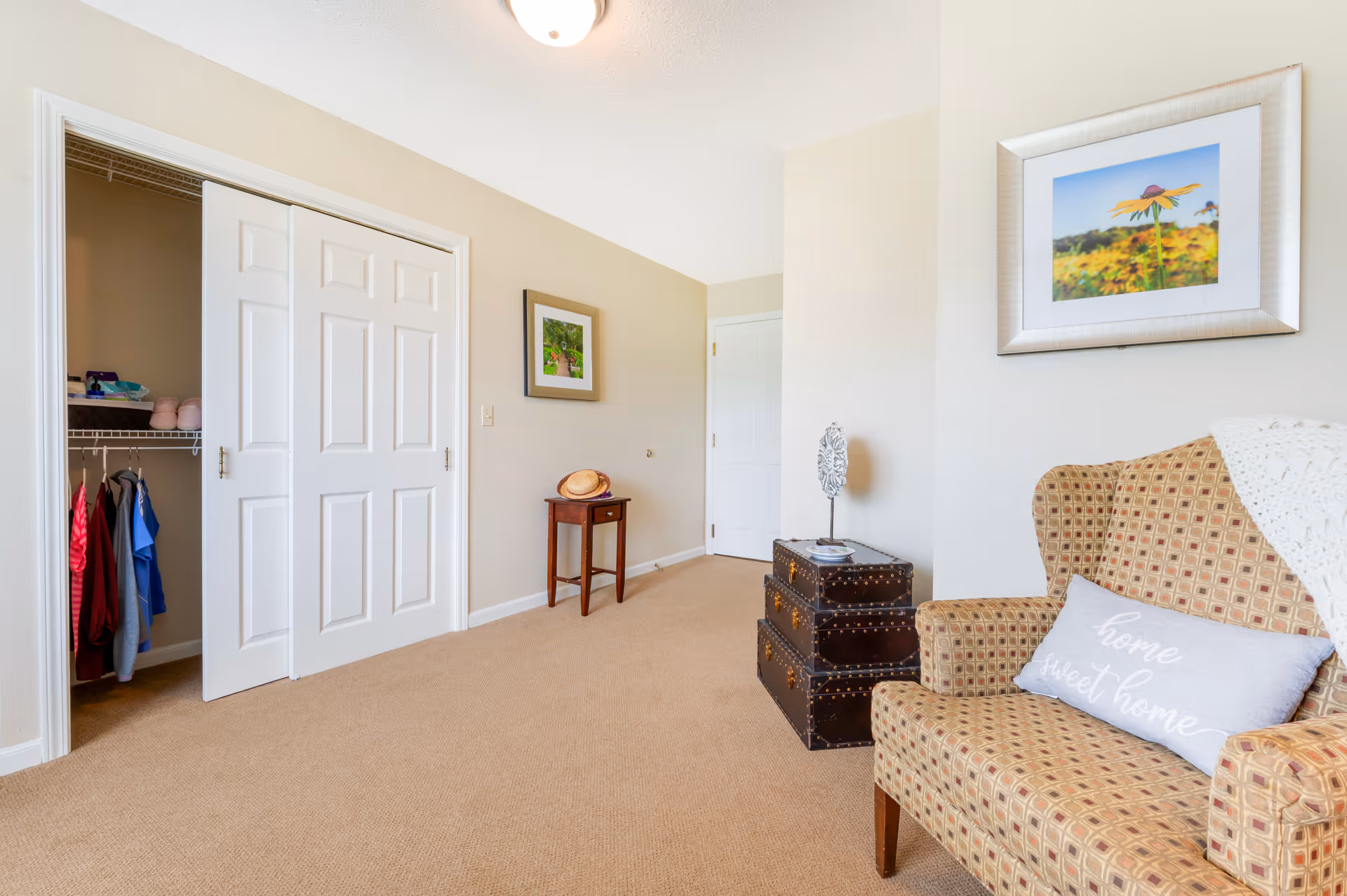 A cozy room with beige carpet and light-colored walls featuring a patterned armchair with a 'home sweet home' pillow, two stacked decorative trunks, a small wooden side table with a hat on it, and an open closet with clothes hanging inside. Two framed pictures hang on the walls.