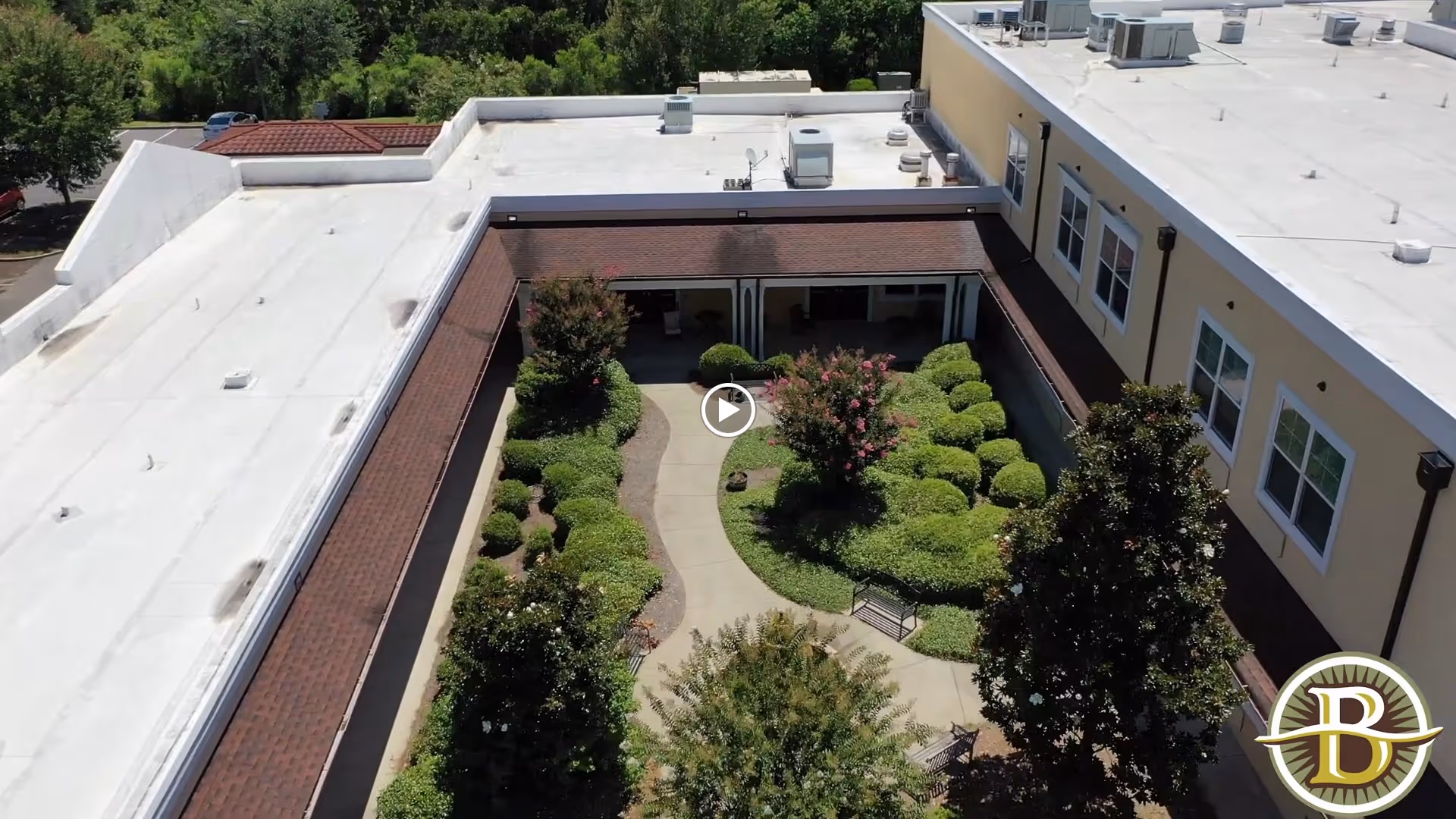 Aerial view of a landscaped courtyard garden within a senior living facility. The courtyard features a winding concrete pathway, neatly trimmed bushes, flowering trees, and benches for seating. The surrounding building has a white roof and beige walls with multiple windows.