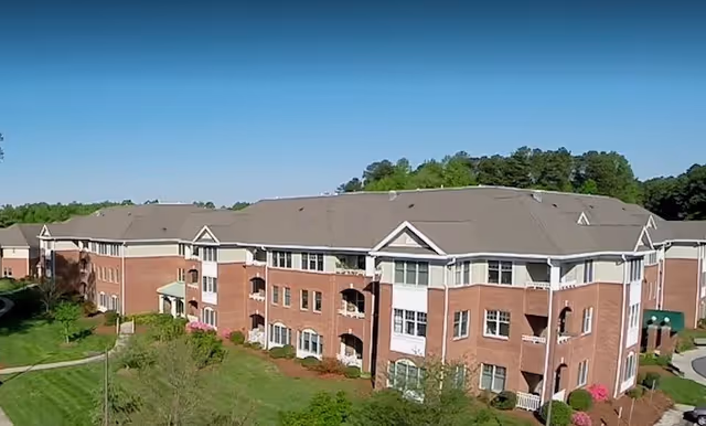 Exterior front view of a large three-story red-brick senior living building with balconies and landscaped lawns.