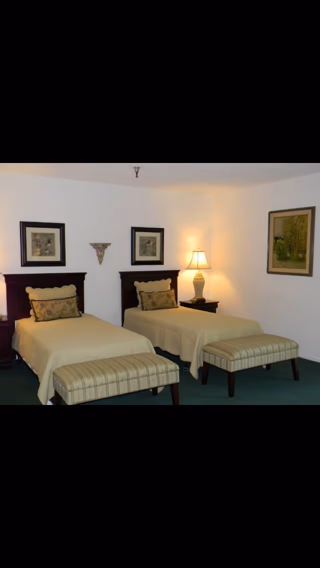 A bedroom with two single beds, each with a beige bedspread and decorative pillows. At the foot of each bed is a striped upholstered bench. A nightstand with a lamp is positioned between the beds. The walls are white and decorated with framed artwork.