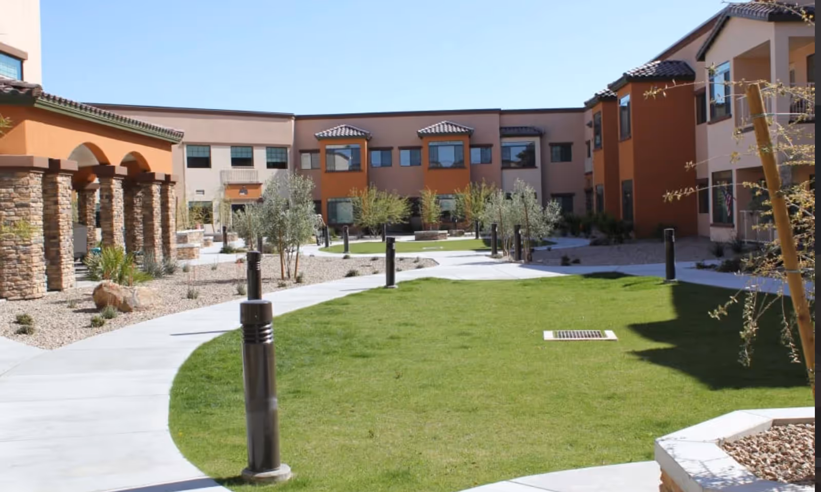 Outdoor courtyard area of Orchard Pointe at Terrazza featuring a green lawn, curved concrete walkways, small trees, and a building with stone pillars and orange and beige walls under a clear blue sky.