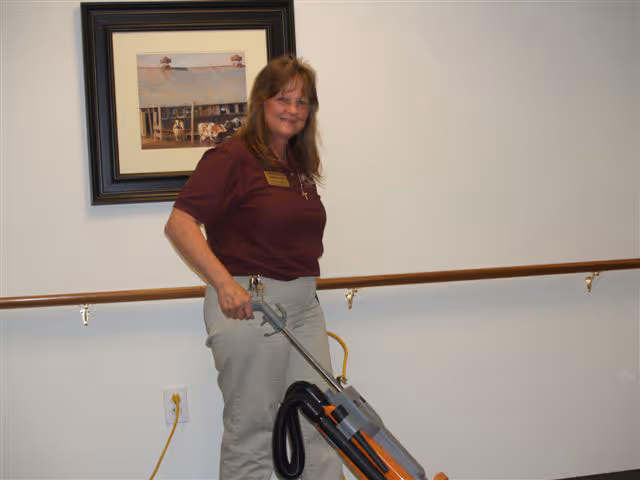A woman with long brown hair wearing a maroon shirt and light-colored pants is vacuuming a carpeted floor in a hallway with a wooden handrail and a framed picture on the wall.