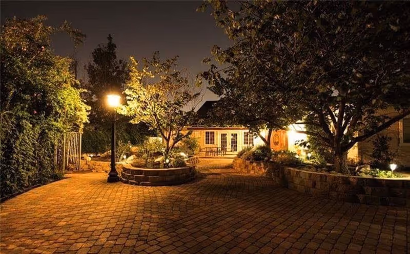 A warmly lit outdoor courtyard at night featuring a paved brick pathway, trees, and a building with large windows and a door in the background. The area is illuminated by a street lamp and garden lights, creating a cozy and inviting atmosphere.