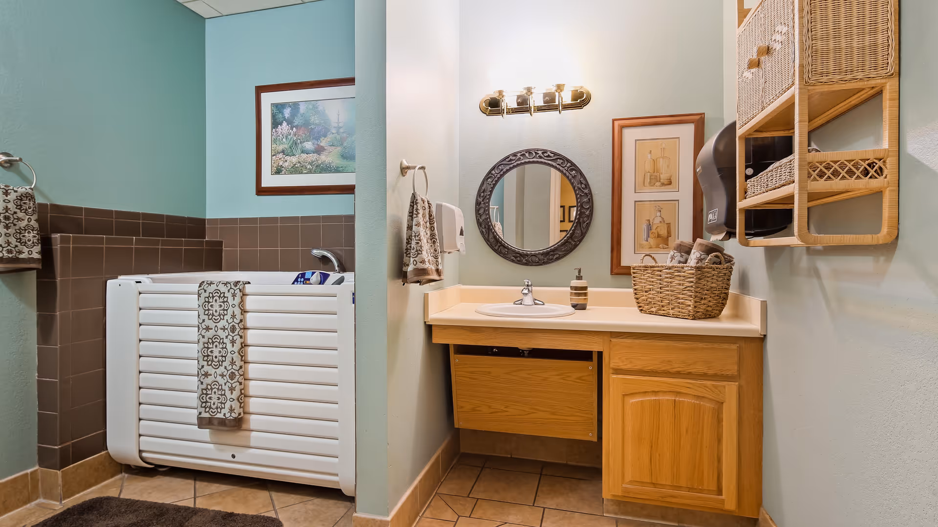 A bathroom with a light blue wall and brown tiled lower half. There is a white accessible bathtub with a patterned towel hanging on its side. Next to the tub is a wooden vanity with a sink, a round decorative mirror above it, and a basket with rolled towels. A wall-mounted paper towel dispenser and a wicker shelf with baskets are also visible. The floor is tiled in a beige color.