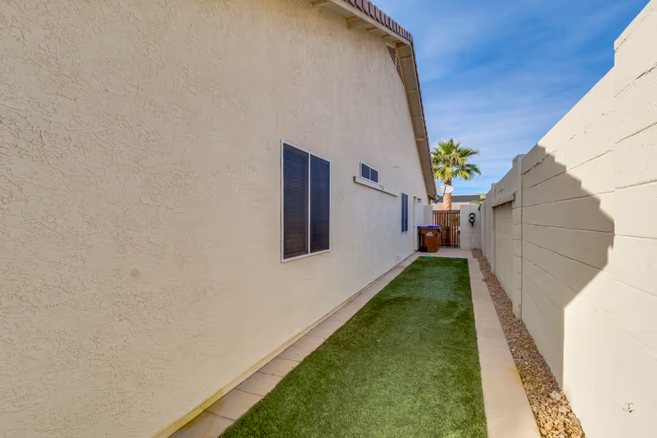 Narrow outdoor side yard with artificial grass strip between a beige stucco building wall and a white block wall, with a brown trash bin and a palm tree visible near a wooden gate at the end under a clear blue sky.