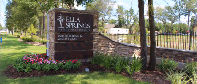 Stone sign for Ella Springs Senior Living Community with the text 'Assisted Living & Memory Care' surrounded by landscaped greenery, flowers, and trees, with a fence and building in the background.