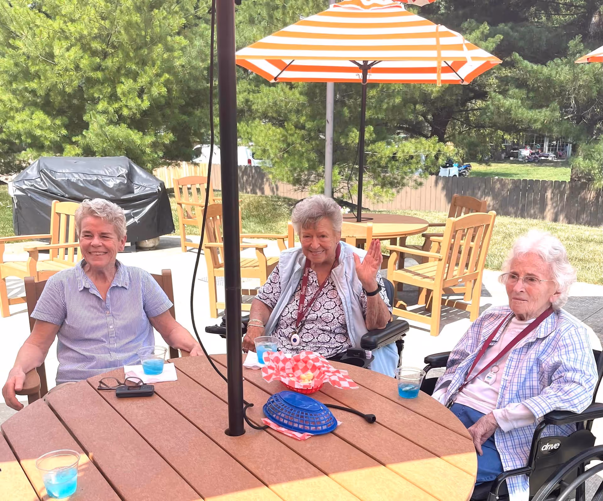 Three elderly women sitting around a round outdoor table with orange and white striped umbrellas. Two women are in wheelchairs, and one woman is seated in a regular chair. They have blue drinks on the table and are enjoying a sunny day in a garden area with trees and wooden chairs in the background.