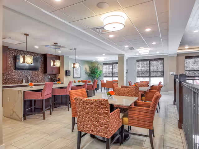 Spacious senior living dining area with tables, patterned upholstered chairs, a bar counter with stools, pendant lights, and large windows.