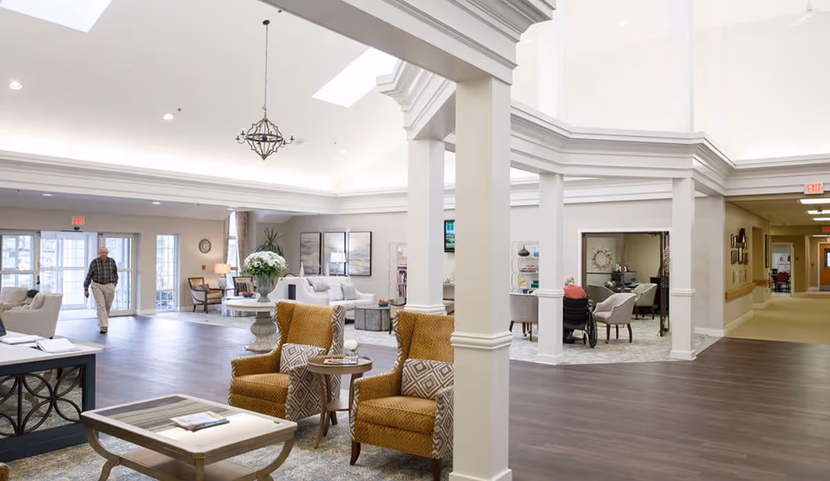 A spacious and well-lit common area in a senior living facility with high ceilings and skylights. The room features comfortable seating including mustard yellow armchairs with patterned cushions, a coffee table, and a round table with a large floral arrangement. In the background, an elderly man walks near large windows and doors, and another elderly person in a wheelchair is seated near a small table. The decor is neutral with light walls, dark wood flooring, and elegant architectural details.