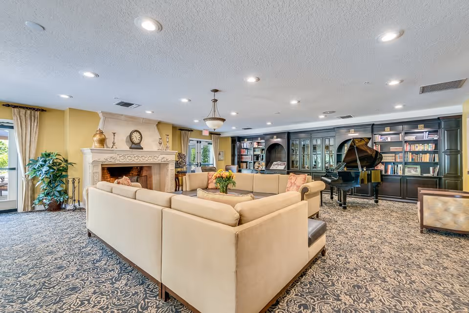 A spacious and well-lit living room area in a senior living facility featuring a large beige sectional sofa arranged around a central coffee table with a flower arrangement. The room includes a decorative fireplace with a clock and vases on the mantel, a grand piano, and built-in dark wood bookshelves filled with books. The carpet has a detailed pattern, and there are large windows with curtains allowing natural light to enter.