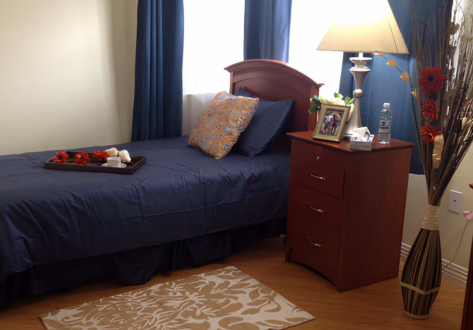 A neatly made single bed with dark blue bedding and two pillows, one blue and one patterned, next to a wooden nightstand with a lamp, a framed photo, a water bottle, and a box of tissues. There is a decorative vase with dried flowers beside the nightstand and a beige patterned rug on the wooden floor.