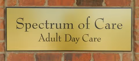 A rectangular sign mounted on a brick wall that reads 'Spectrum of Care Adult Day Care' in black text on a yellow background.