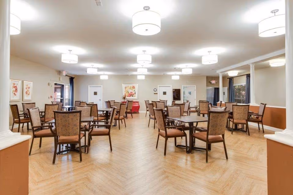 A spacious dining room with multiple square tables and cushioned chairs arranged neatly. The room has light-colored walls, wooden flooring, several ceiling lights, framed artwork on the walls, and windows with dark curtains.