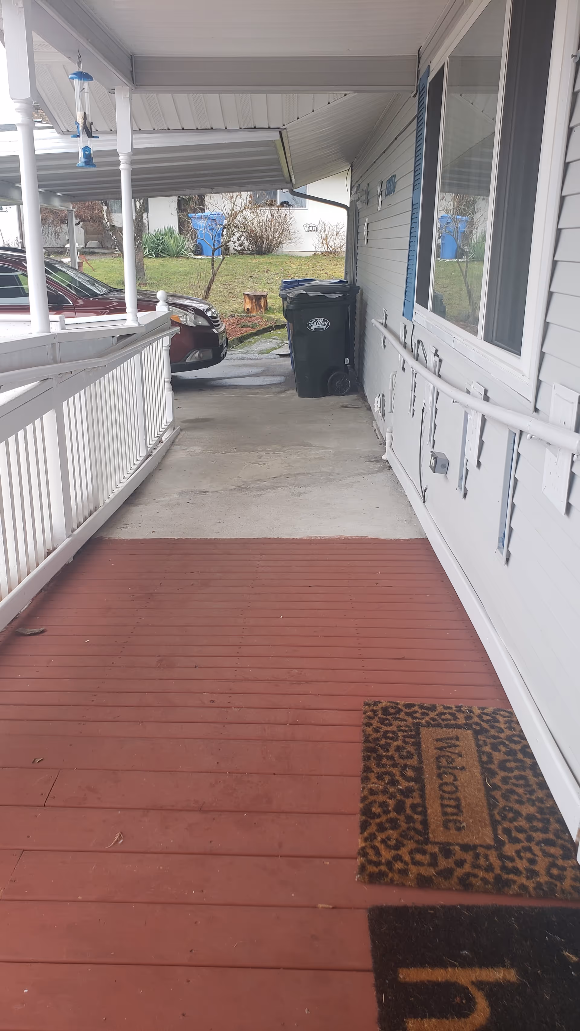 Covered front porch and driveway with white railing, red painted floorboards, a welcome mat, a trash bin, and a parked car visible.