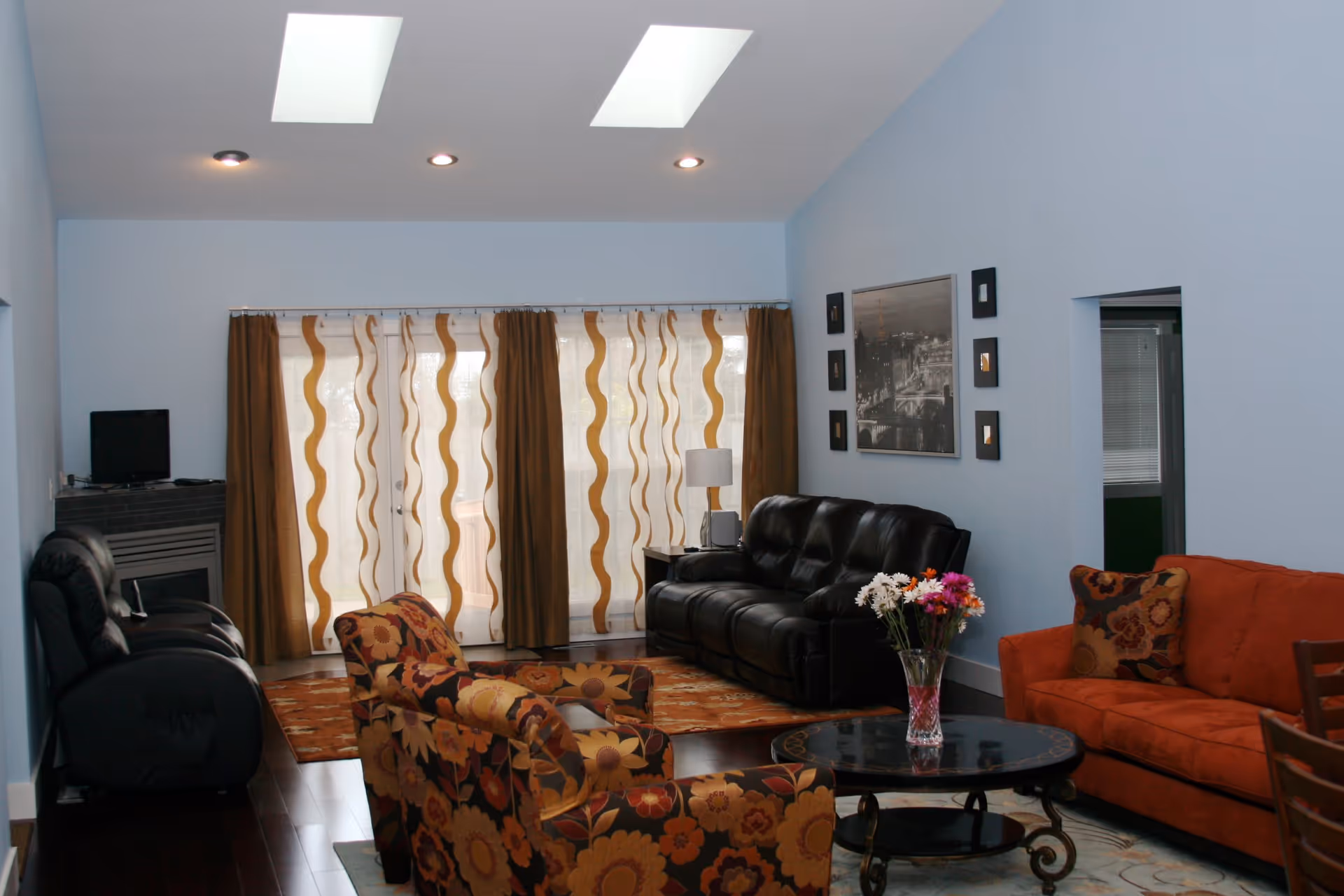A living room with light blue walls and dark wooden floors. The room features a black leather sofa, an orange sofa with a floral cushion, and two floral-patterned armchairs. There is a round black coffee table with a vase of colorful flowers on it. The back wall has large windows covered with sheer curtains with a wavy pattern and brown drapes. The ceiling has two skylights and recessed lighting. A black and white framed picture and smaller framed art pieces hang on the wall above the black sofa.