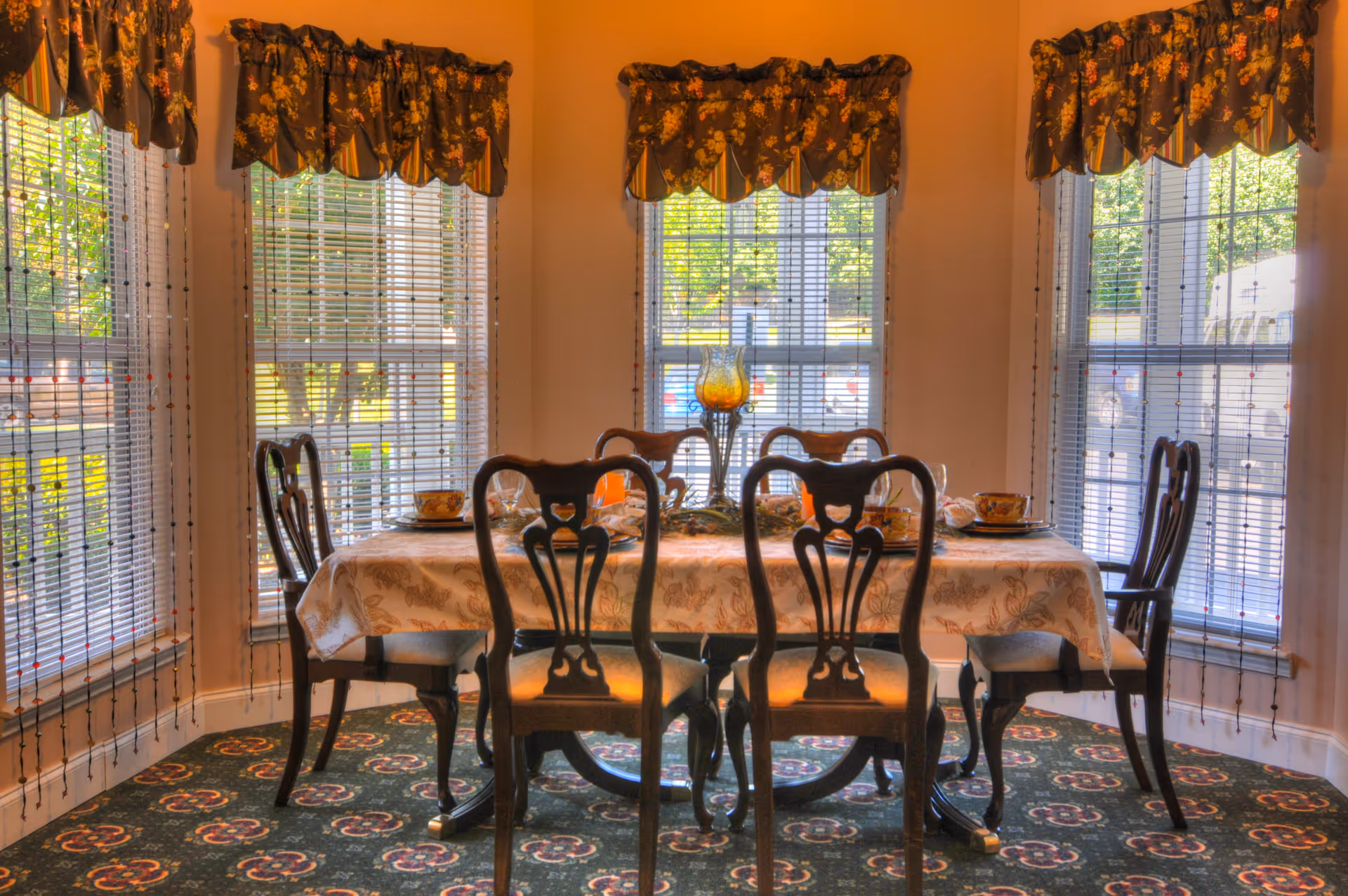A formal dining room with a table set for six in front of large bay windows with floral valances.